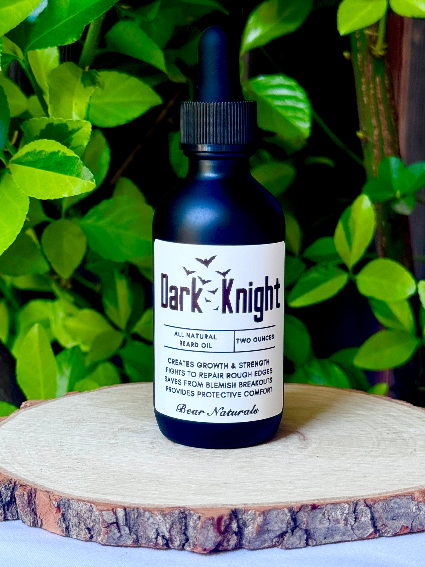 Two ounce black bottle of Dark Knight all-natural beard oil on a wooden surface with green leaves in the background