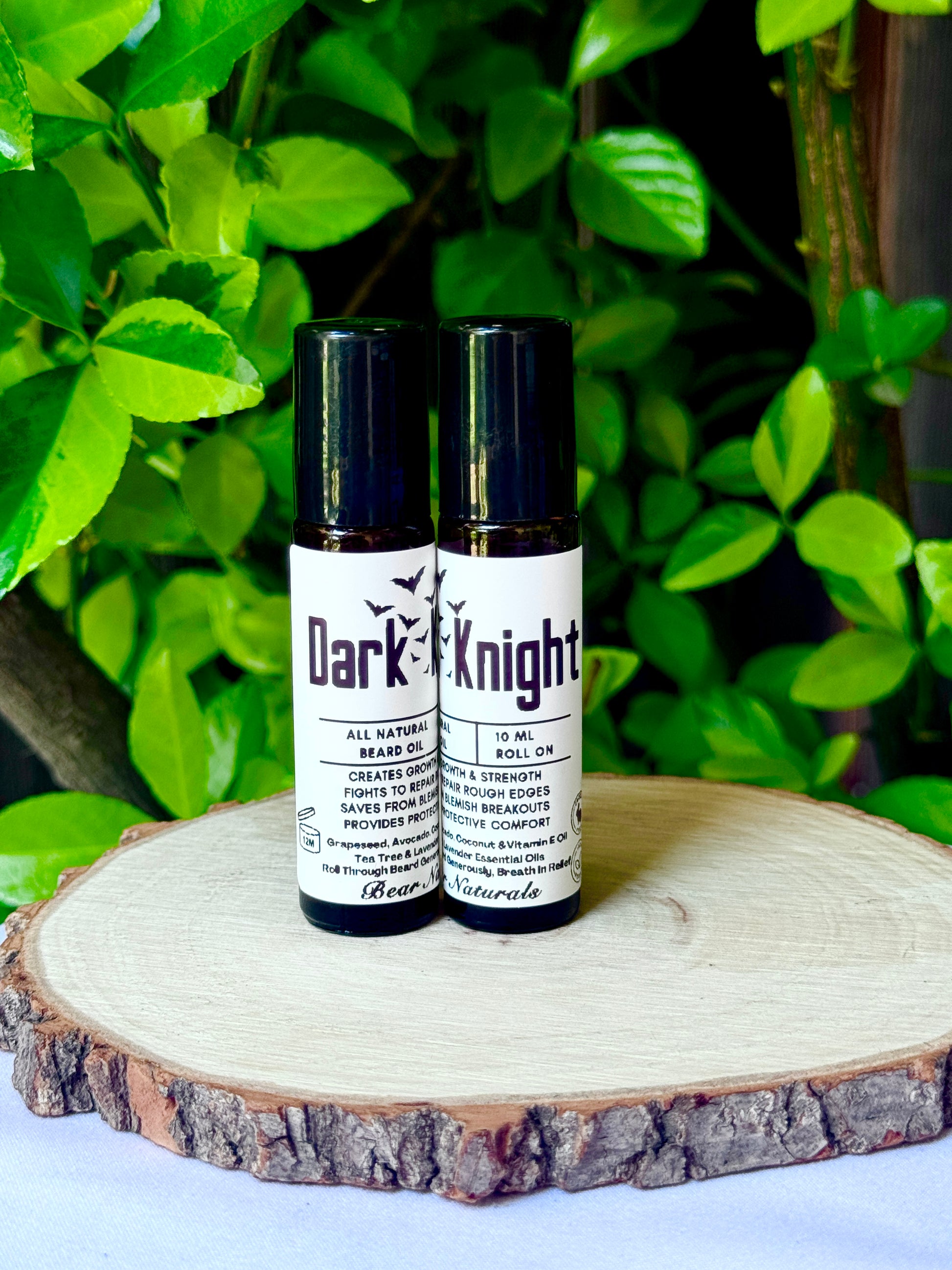 Two bottles of 'Dark Knight' all-natural beard oil on a wooden surface with green leaves in the background.
