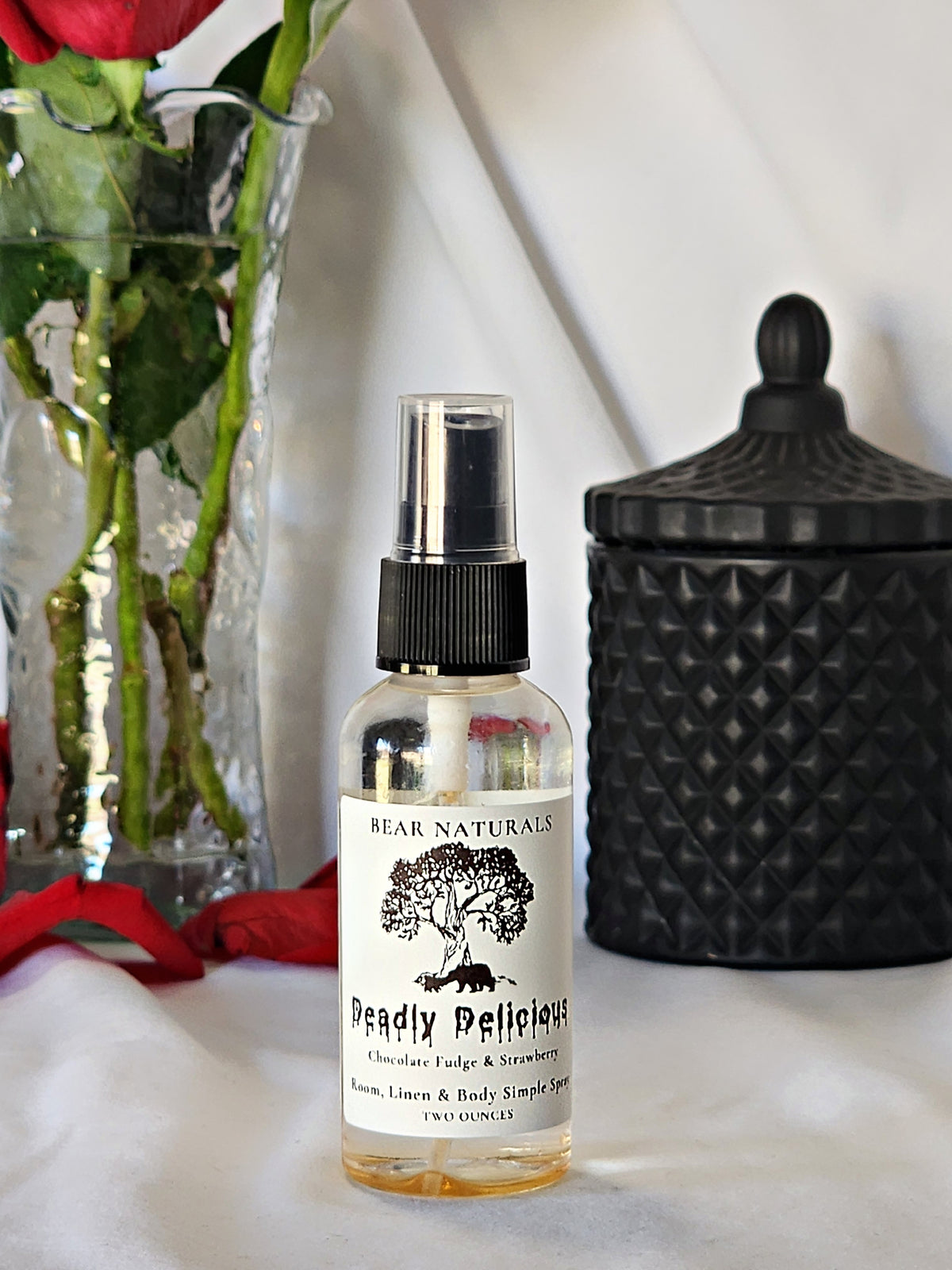 A clear bottle with a black fingertip style spray nozzle featuring a white label with black writing with a black candle jar and red roses in a vase in the background.