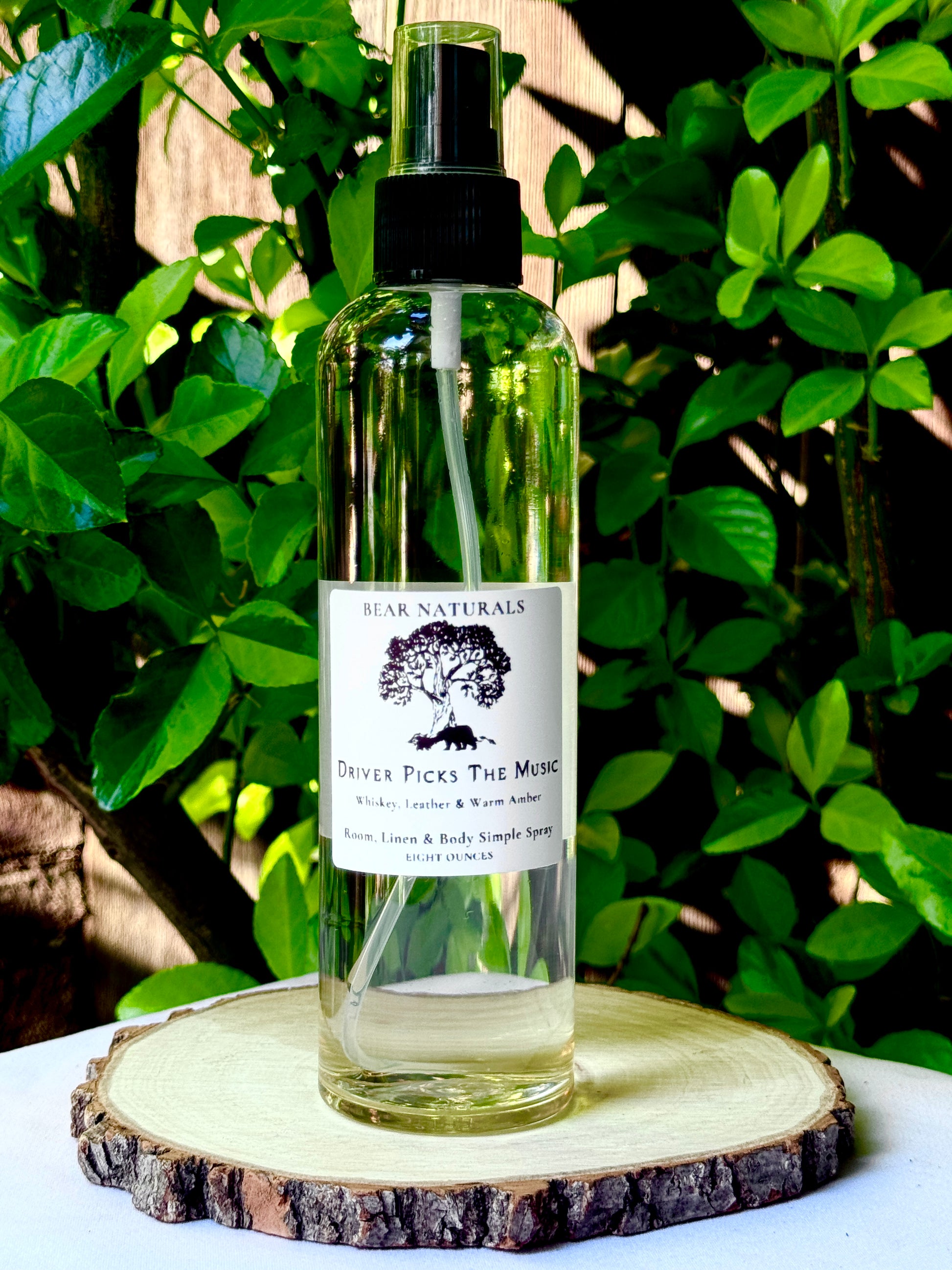 A clear bottle with a black fingertip style spray nozzle featuring a white label with black writing on a wooden surface with green leaves in the background.