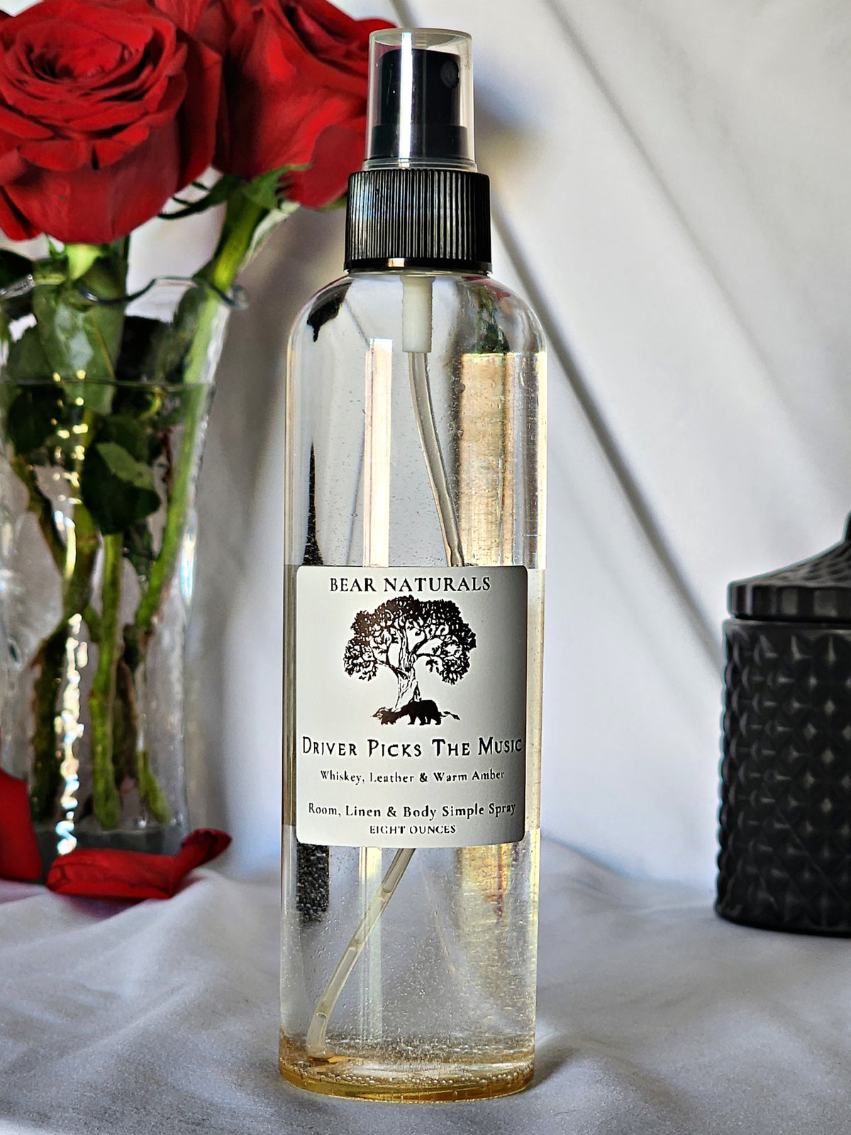 A clear bottle with a black fingertip style spray nozzle featuring a white label with black writing with a black candle jar and red roses in a vase in the background.