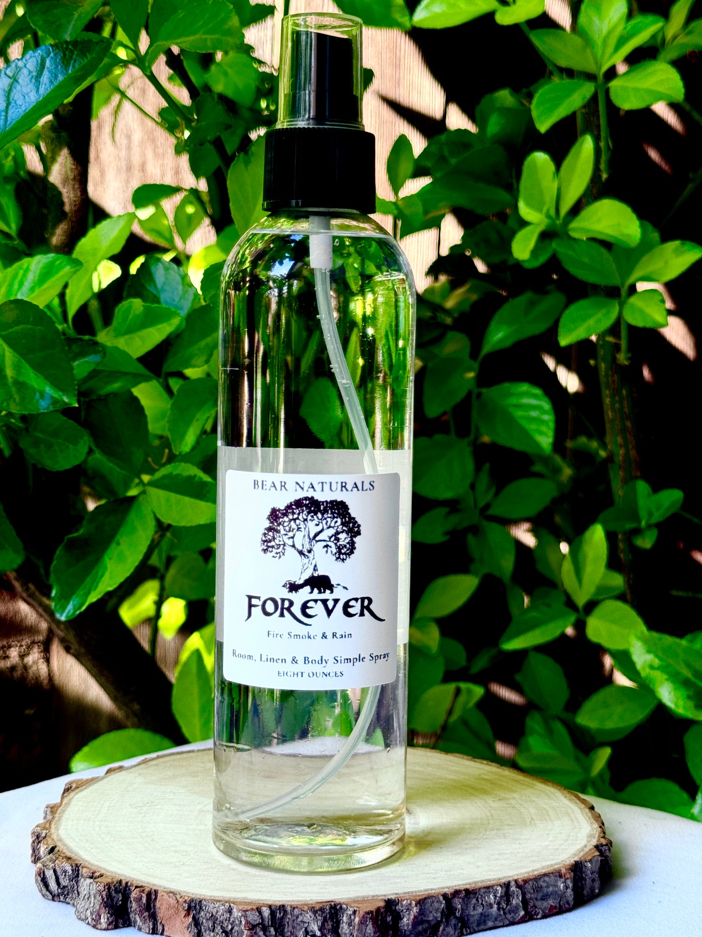 A clear bottle with a black fingertip style spray nozzle featuring a white label with black writing on a wooden surface with green leaves in the background.