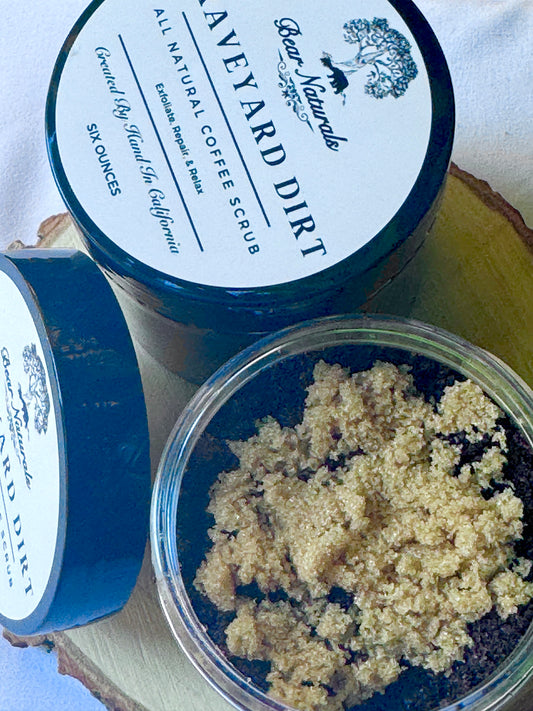 A six ounce clear jar containing exfoliating coffee scrub topped with brown sugar sitting on a wooden surface with green leaves in the background. The white label with black writing is featured on the lid and reads "Graveyard Dirt All Natural Coffee Scrub".