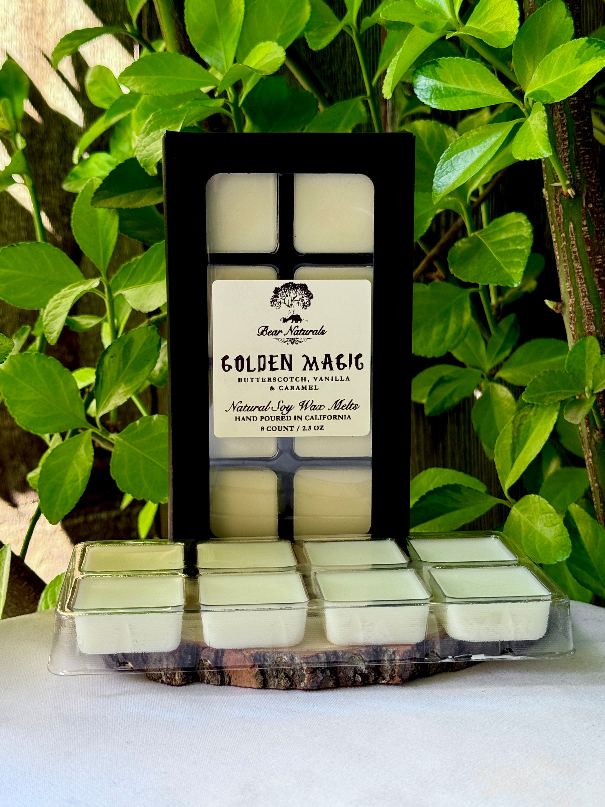 A black box with a clear plastic tray housing eight white wax melts featuring a white label with black writing on a wooden surface with green leaves in the background