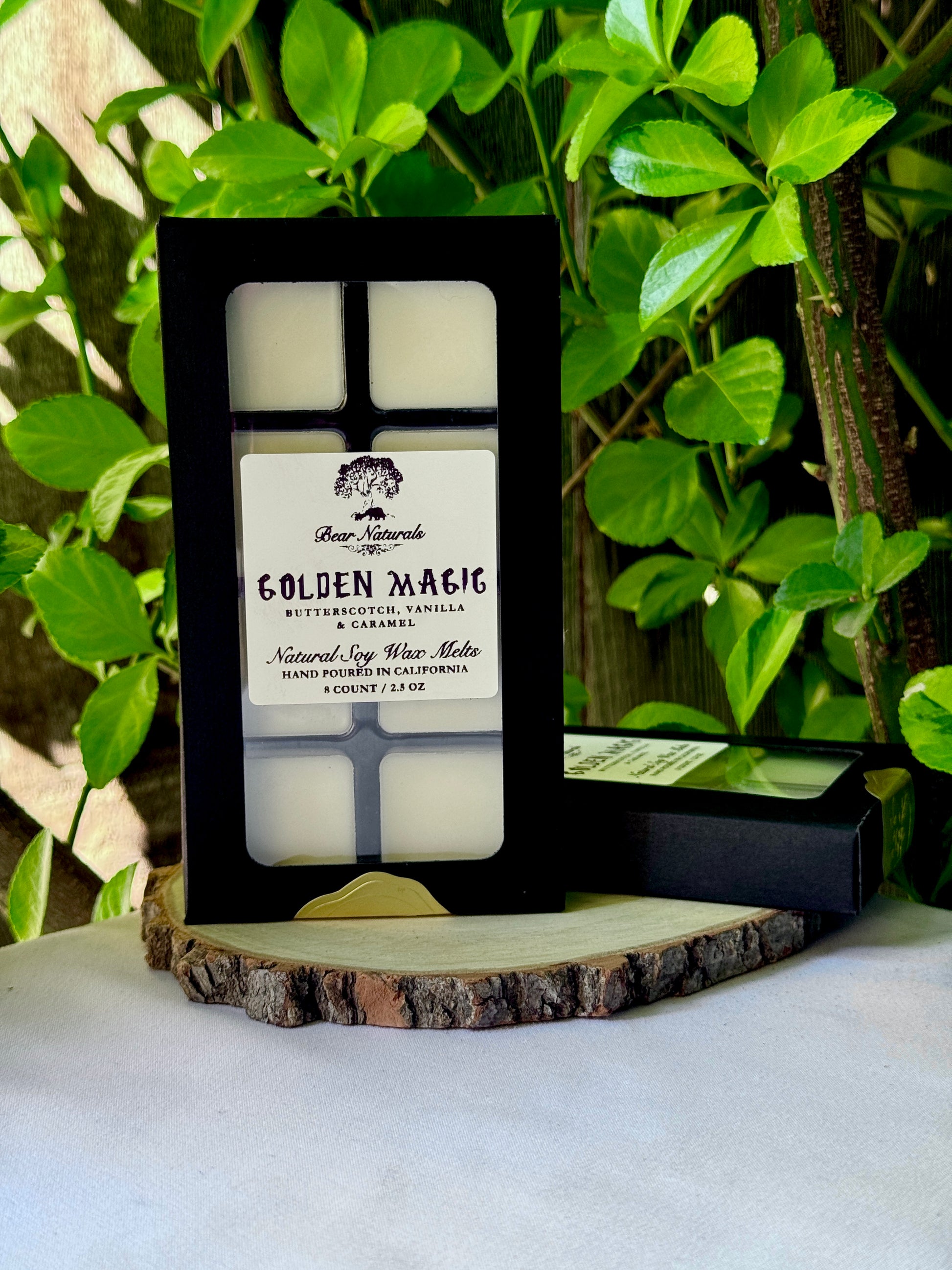 A black box with a clear plastic tray housing eight white wax melts featuring a white label with black writing on a wooden surface with green leaves in the background
