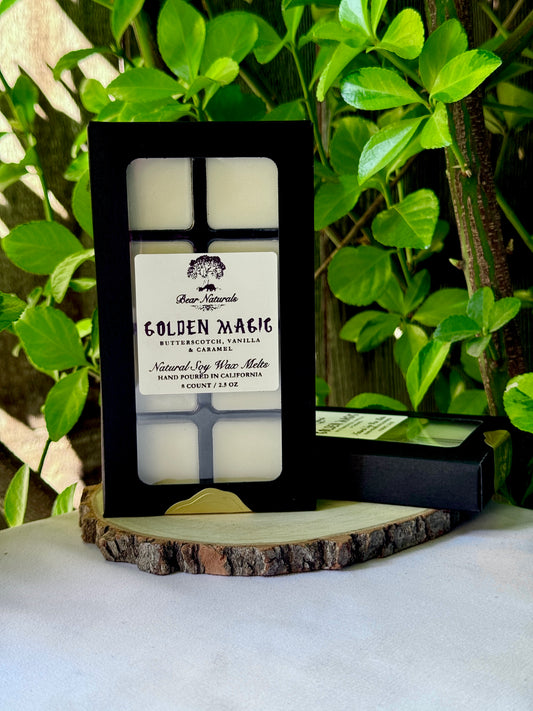 A black box with a clear plastic tray housing eight white wax melts featuring a white label with black writing on a wooden surface with green leaves in the background