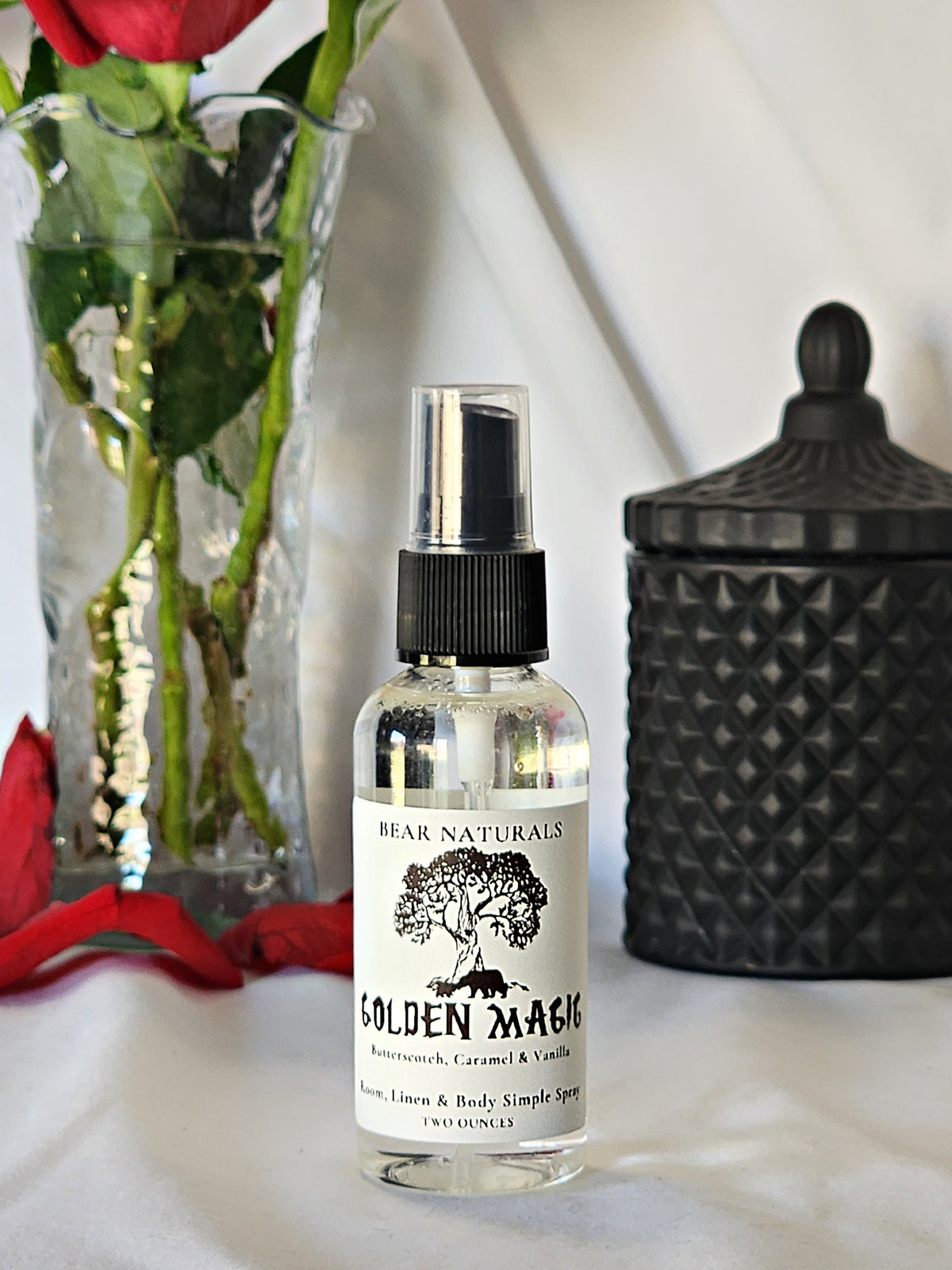 A clear bottle with a black fingertip style spray nozzle featuring a white label with black writing with a black candle jar and red roses in a vase in the background.