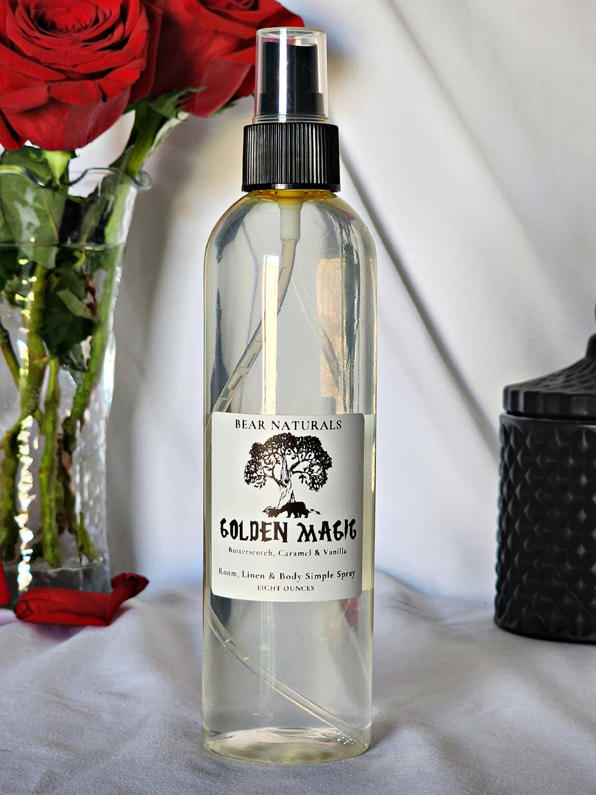 A clear bottle with a black fingertip style spray nozzle featuring a white label with black writing with a black candle jar and red roses in a vase in the background.