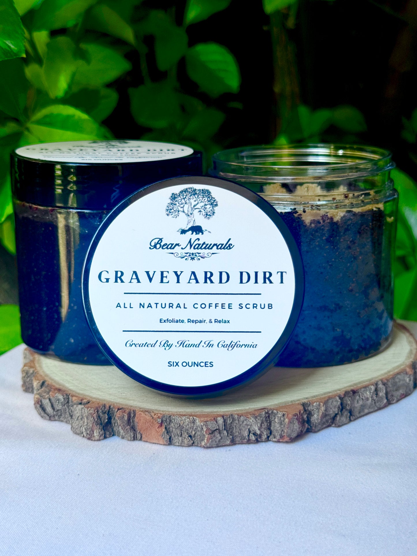 A six ounce clear jar containing exfoliating coffee scrub topped with brown sugar sitting on a wooden surface with green leaves in the background. The white label with black writing is featured on the lid and reads "Graveyard Dirt All Natural Coffee Scrub".