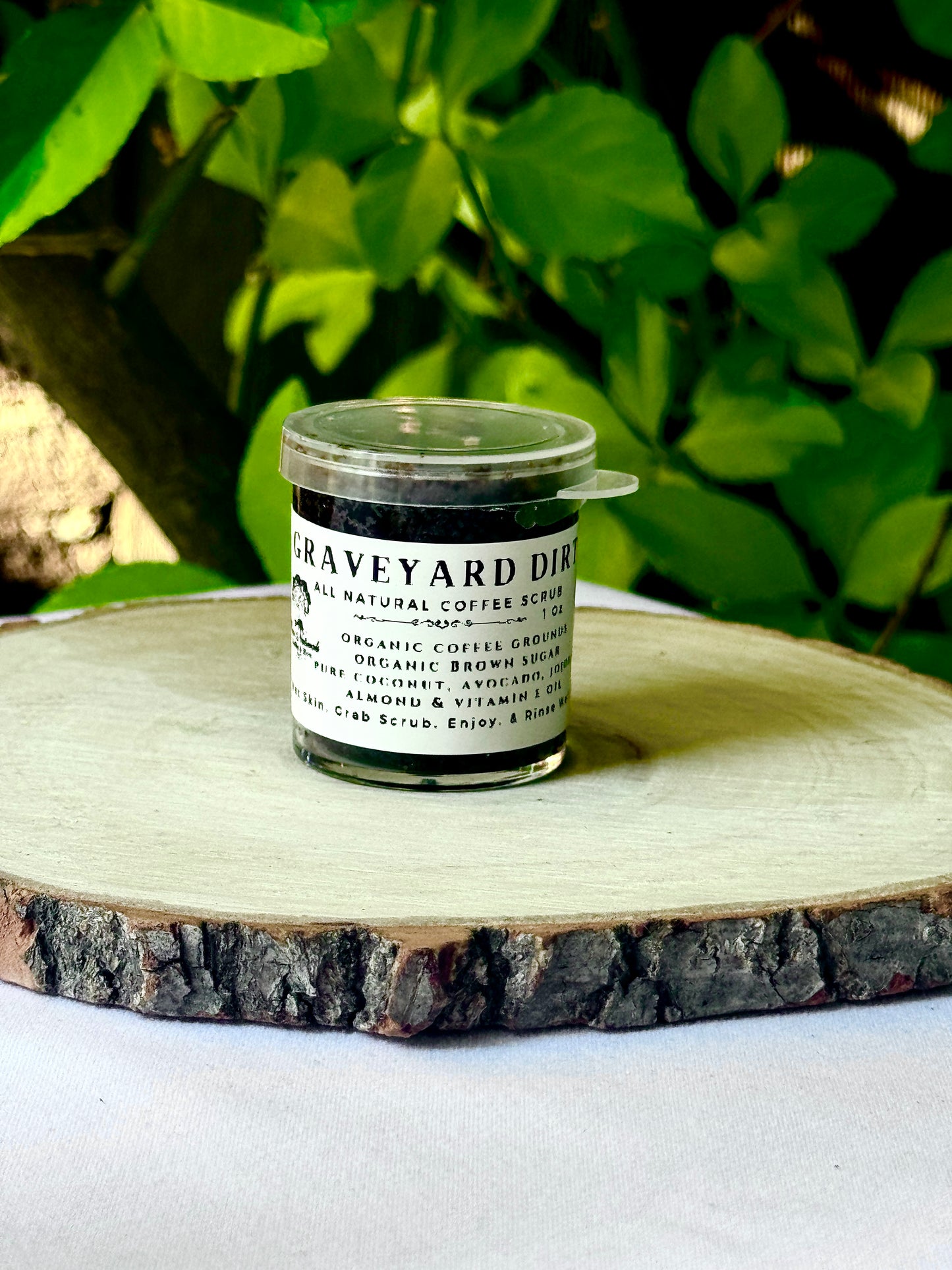 A small clear jar with a clear lid white label with black writing filled with Resuscitate All Natural Exfoliating Sugar Scrub on a wooden surface with green leaves in the background