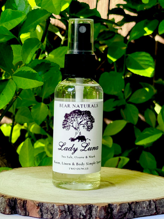 A clear bottle with a black fingertip style spray nozzle featuring a white label with black writing on a wooden surface with green leaves in the background.