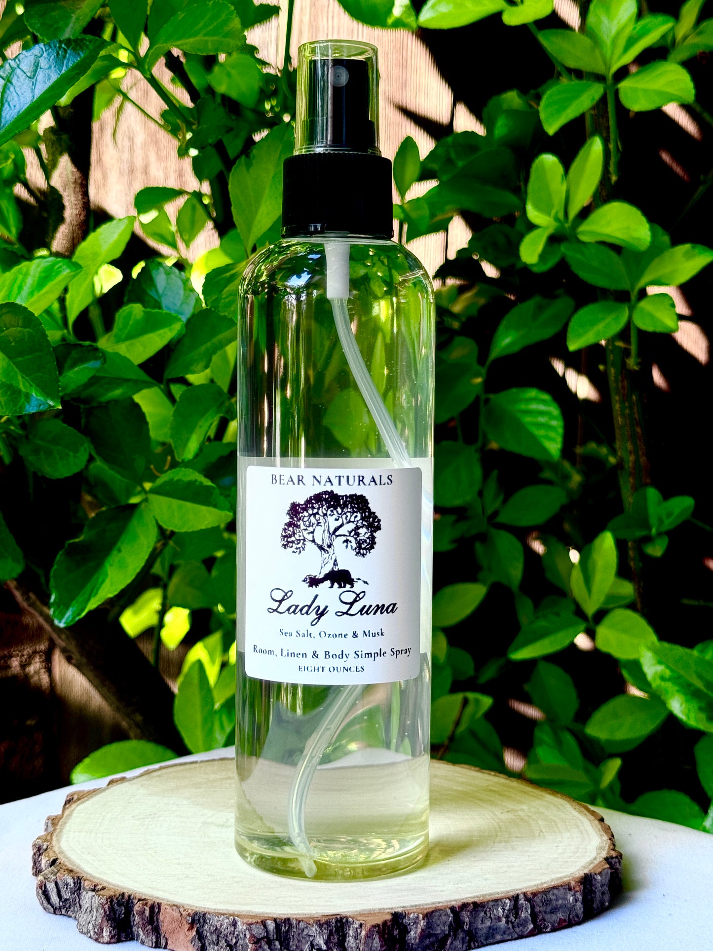 A clear bottle with a black fingertip style spray nozzle featuring a white label with black writing on a wooden surface with green leaves in the background.