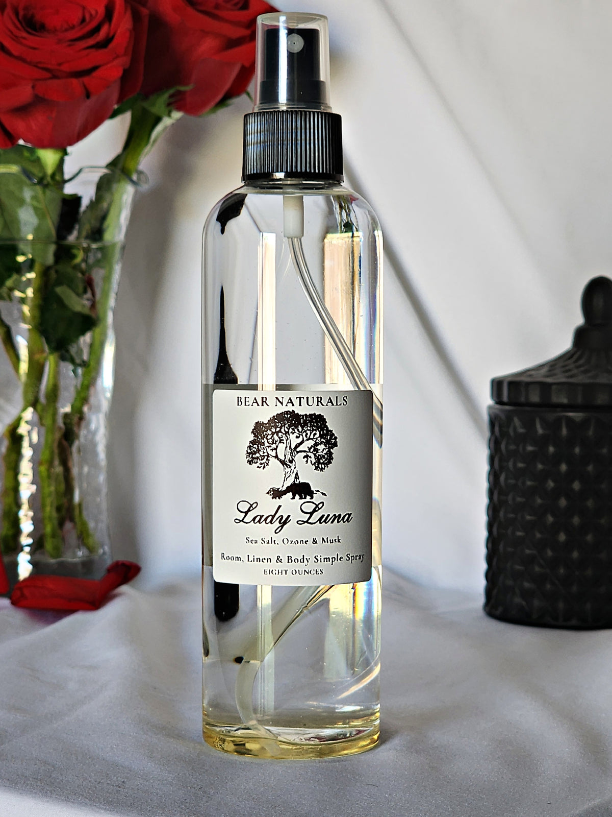 A clear bottle with a black fingertip style spray nozzle featuring a white label with black writing with a black candle jar and red roses in a vase in the background.