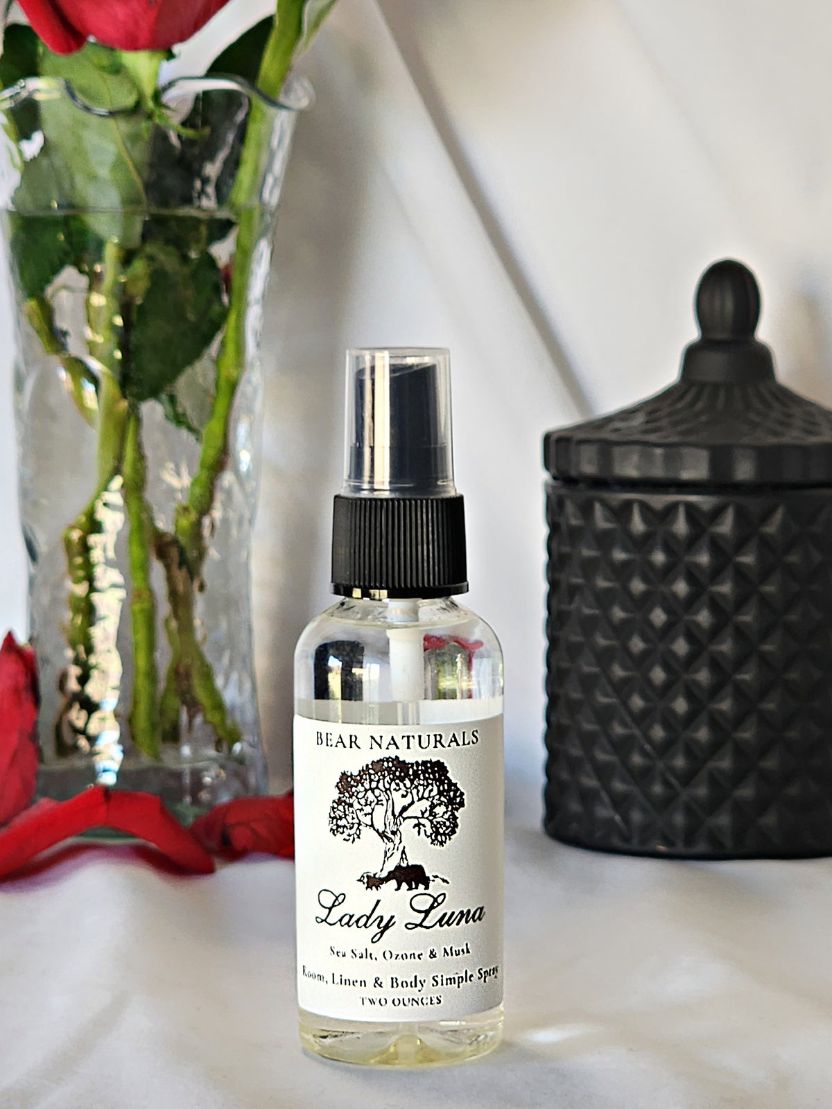 A clear bottle with a black fingertip style spray nozzle featuring a white label with black writing with a black candle jar and red roses in a vase in the background.