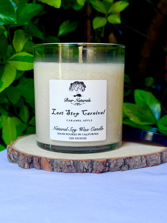 a ten ounce white candle in a clear jar featuring a white label with black writing on a wooden surface with green leaves in the back ground.