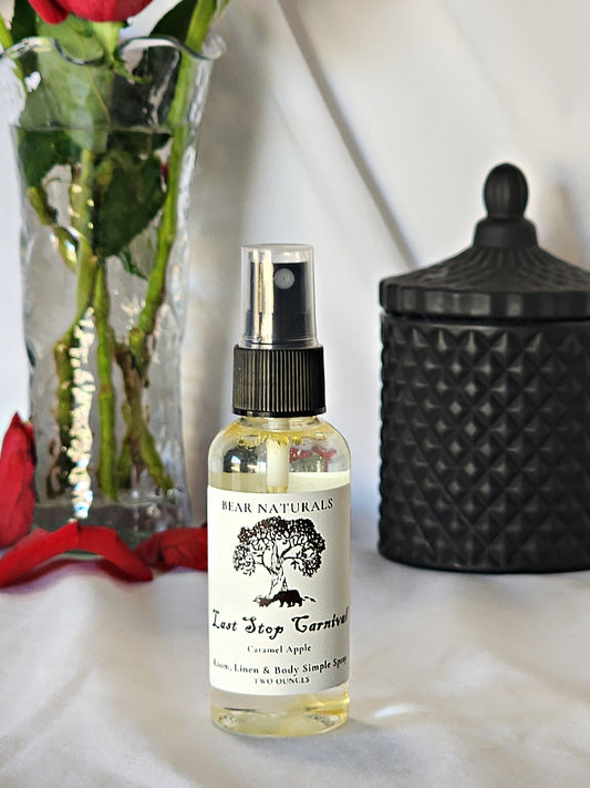 A clear bottle with a black fingertip style spray nozzle featuring a white label with black writing with a black candle jar and red roses in a vase in the background.