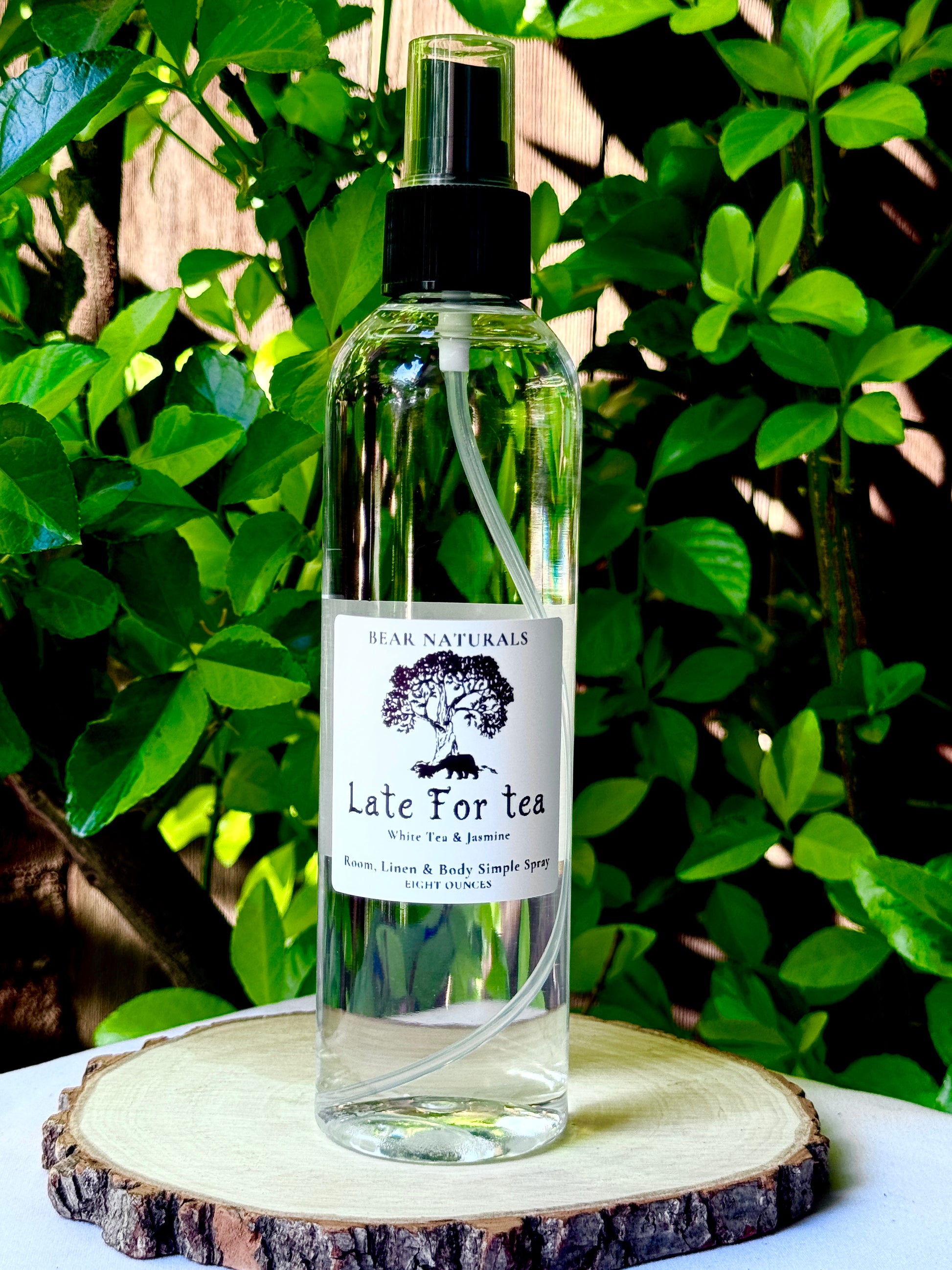 A clear bottle with a black fingertip style spray nozzle featuring a white label with black writing on a wooden surface with green leaves in the background.