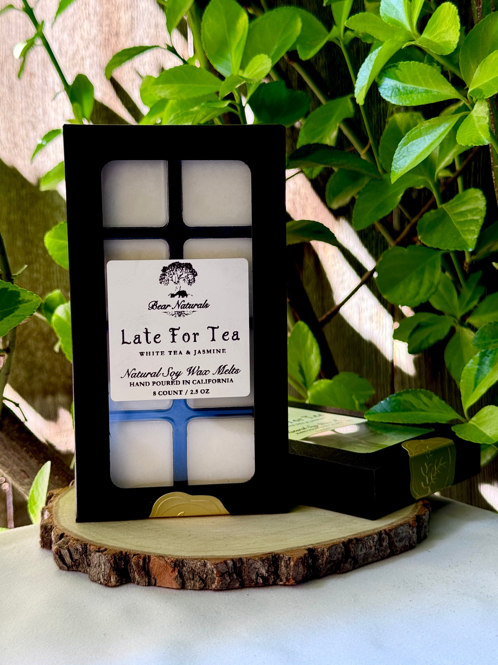 A black box with a clear plastic tray housing eight white wax melts featuring a white label with black writing on a wooden surface with green leaves in the background