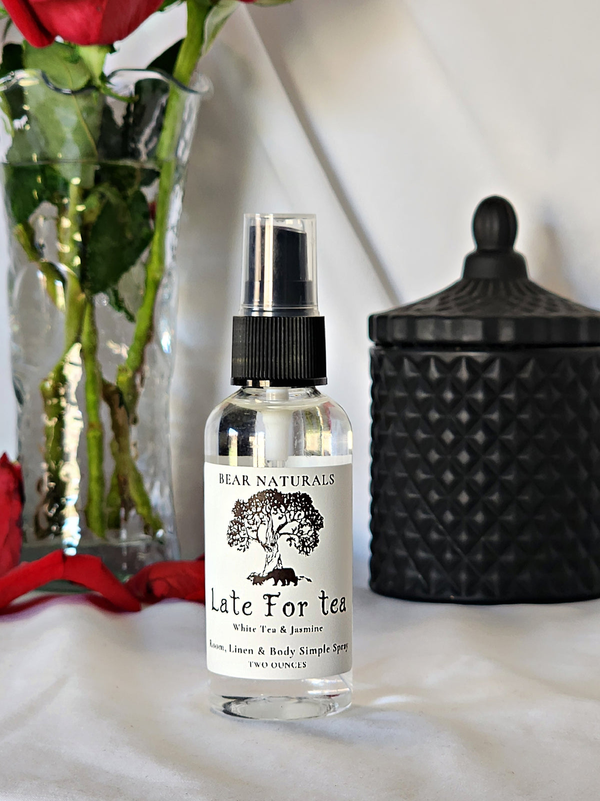 A clear bottle with a black fingertip style spray nozzle featuring a white label with black writing with a black candle jar and red roses in a vase in the background.