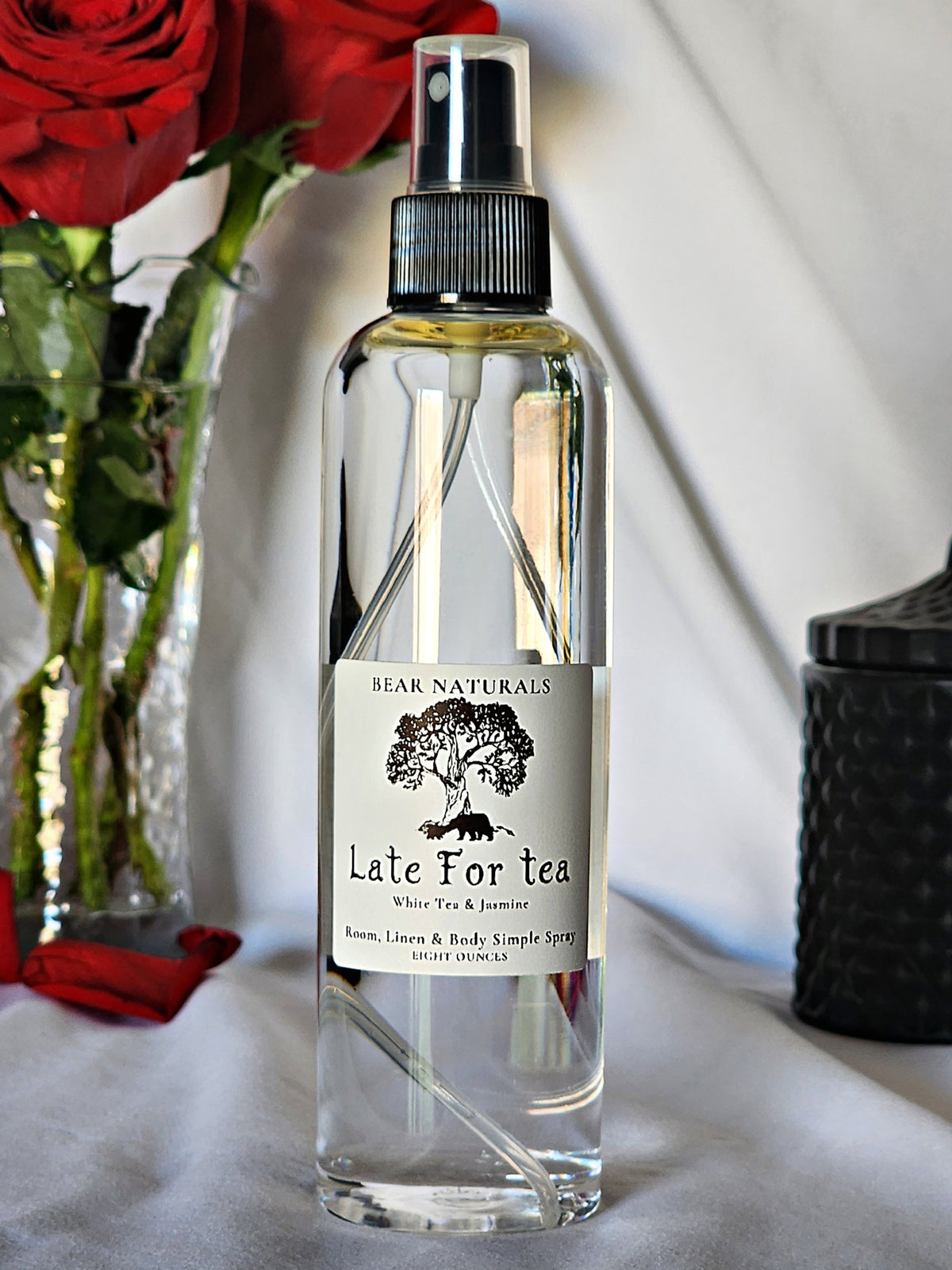 A clear bottle with a black fingertip style spray nozzle featuring a white label with black writing with a black candle jar and red roses in a vase in the background.
