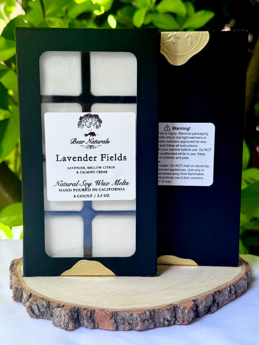 A black box with a clear plastic tray housing eight white wax melts featuring a white label with black writing on a wooden surface with green leaves in the background