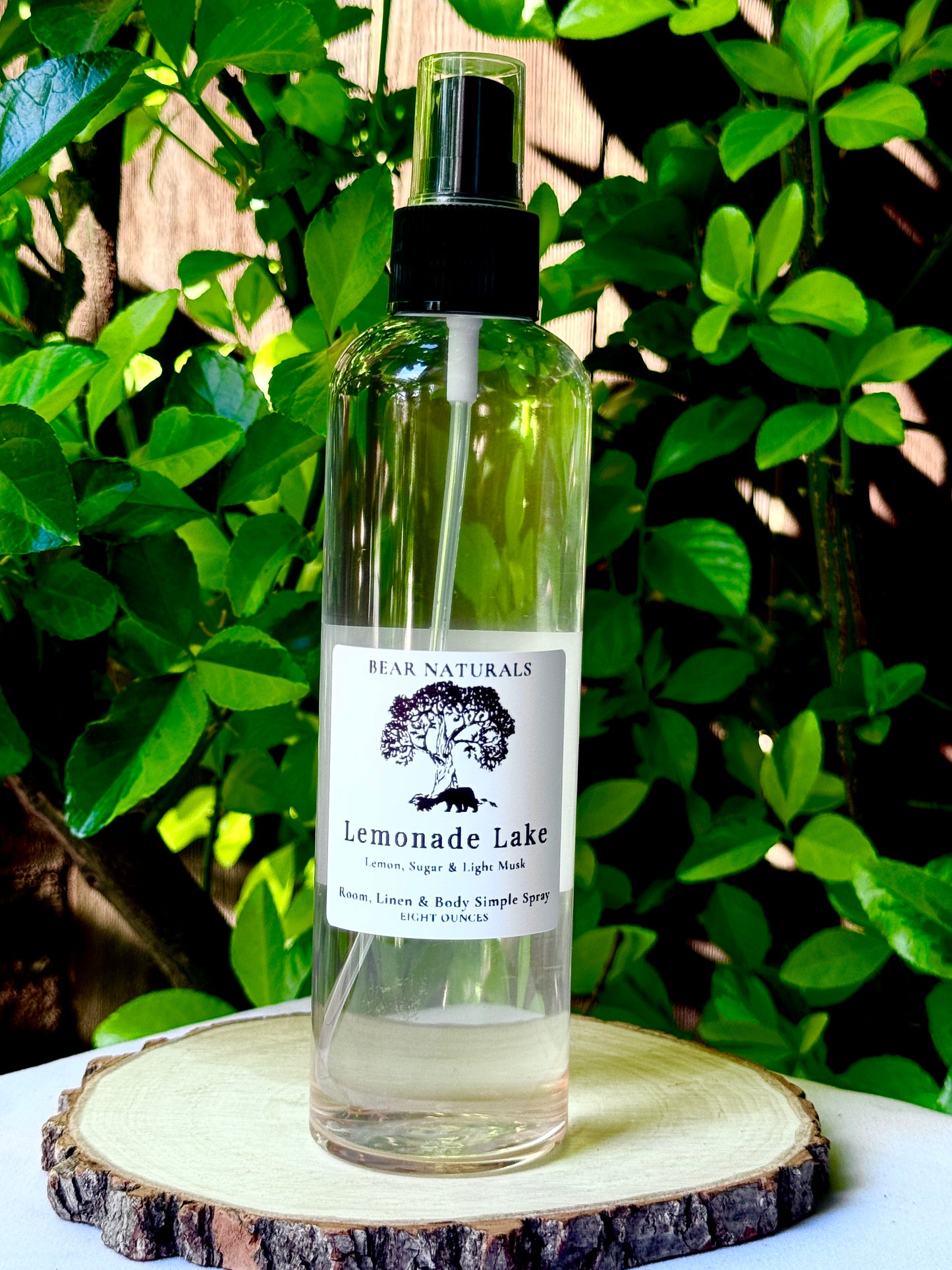 A clear bottle with a black fingertip style spray nozzle featuring a white label with black writing on a wooden surface with green leaves in the background.