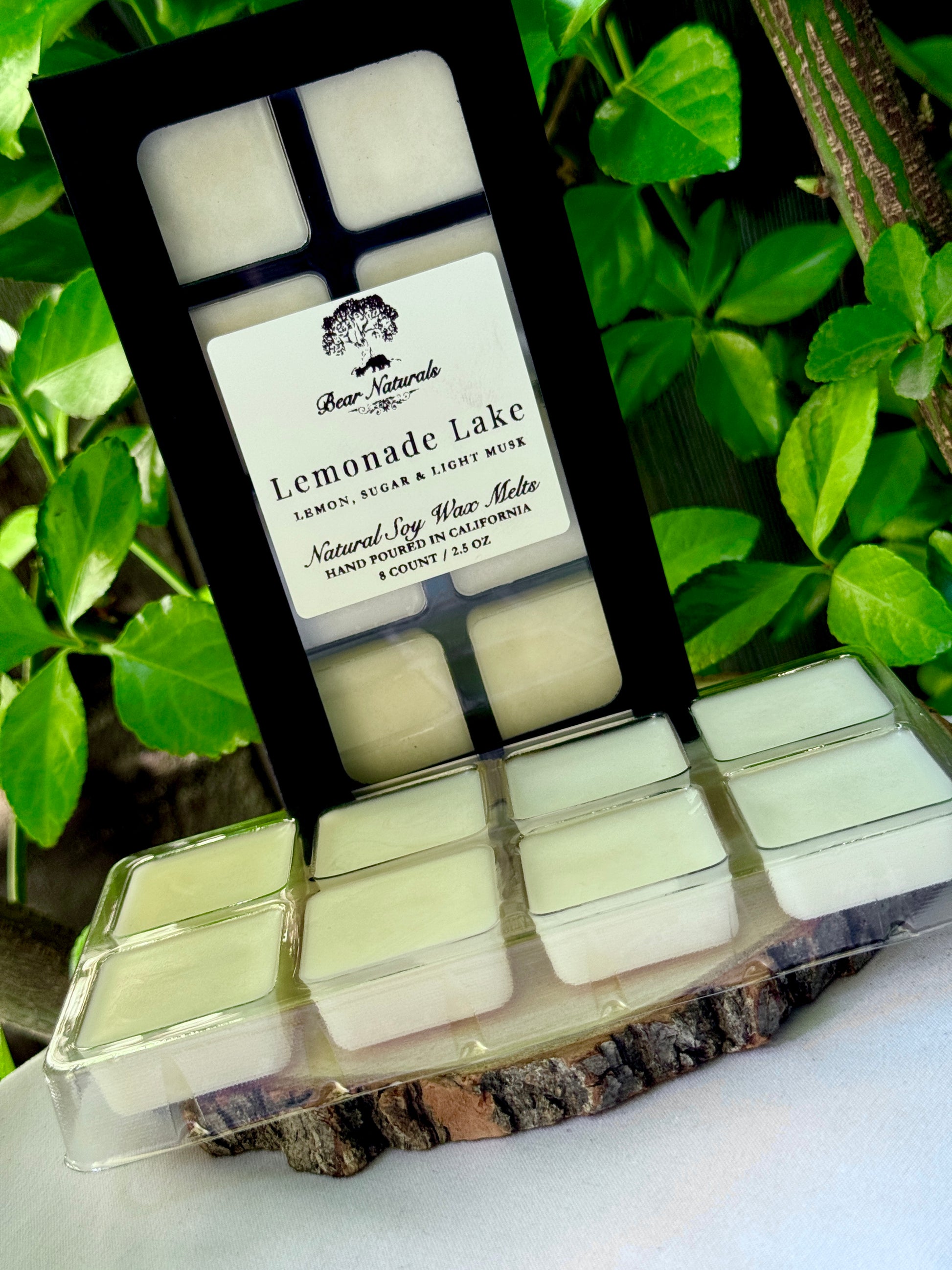A black box with a clear plastic tray housing eight white wax melts featuring a white label with black writing on a wooden surface with green leaves in the background