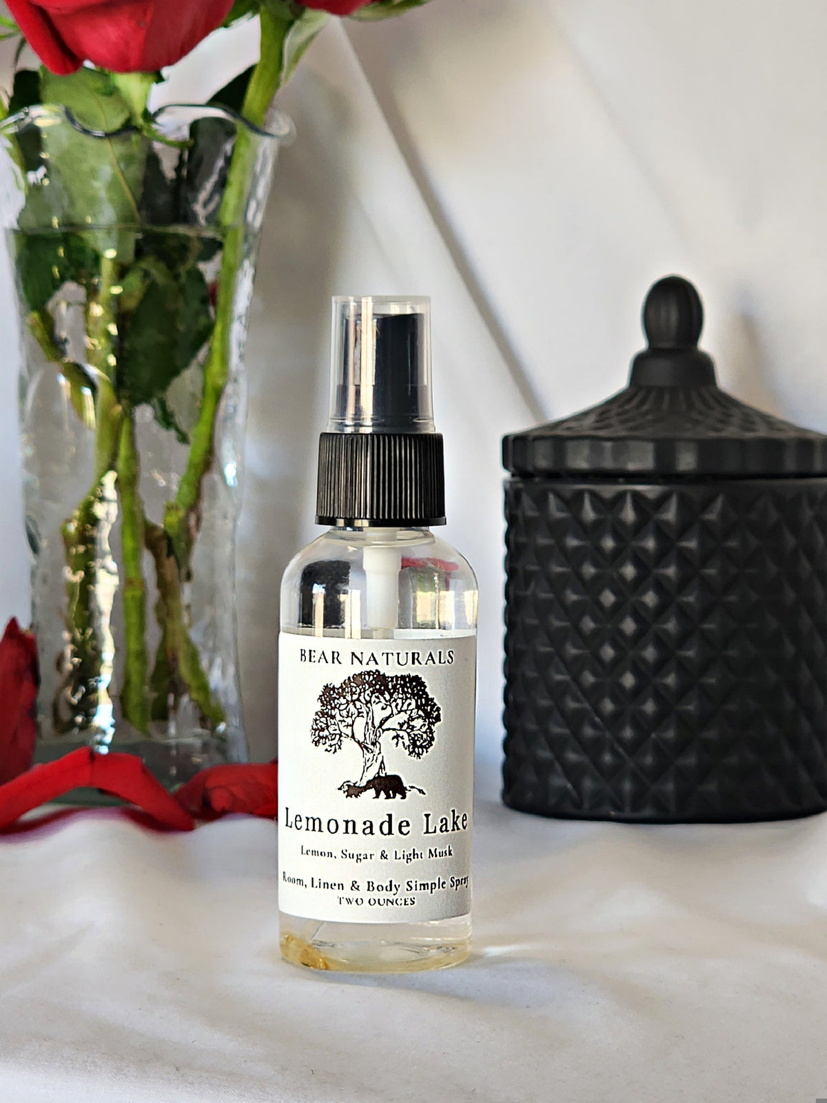 A clear bottle with a black fingertip style spray nozzle featuring a white label with black writing with a black candle jar and red roses in a vase in the background.