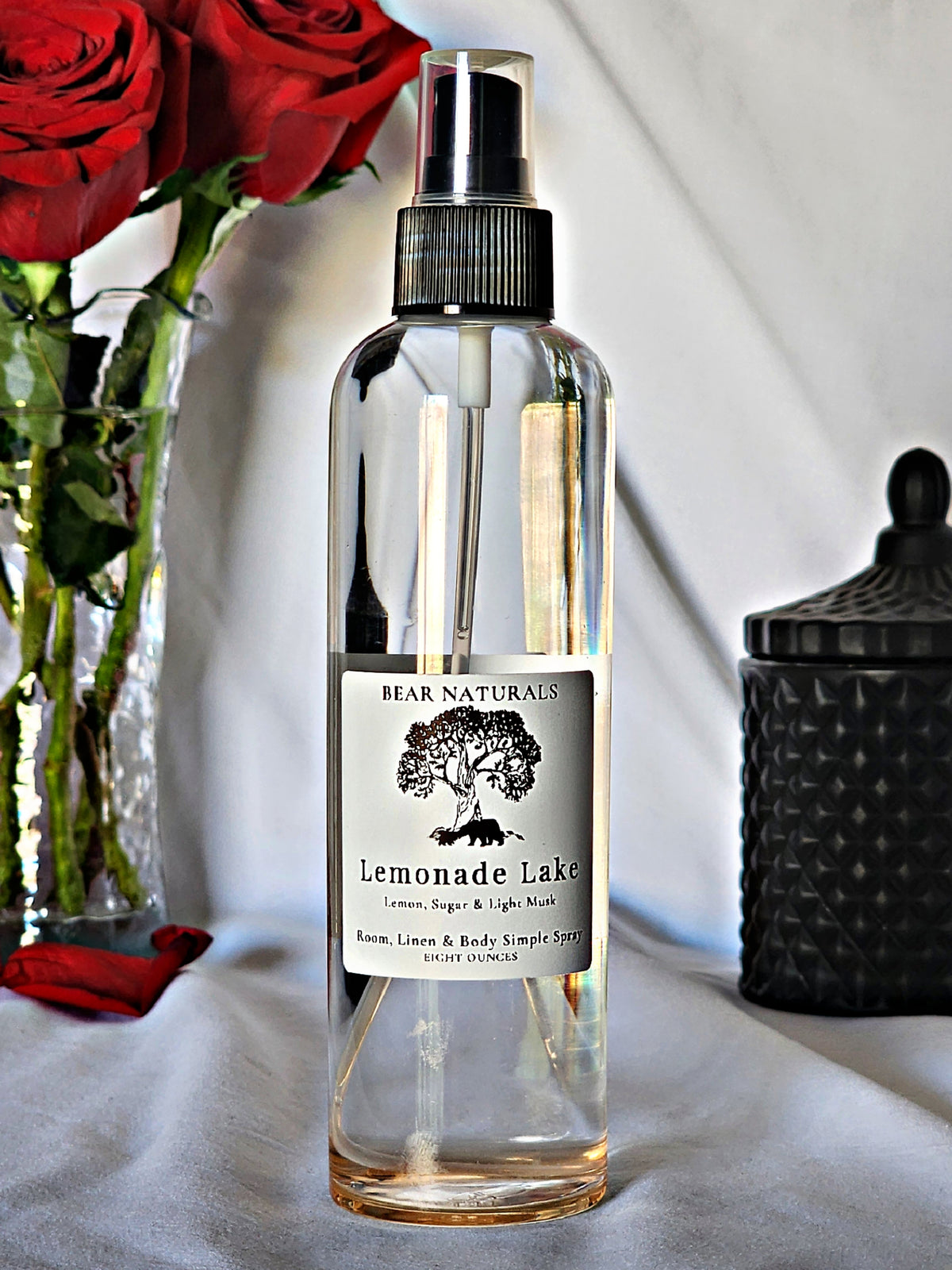 A clear bottle with a black fingertip style spray nozzle featuring a white label with black writing with a black candle jar and red roses in a vase in the background.