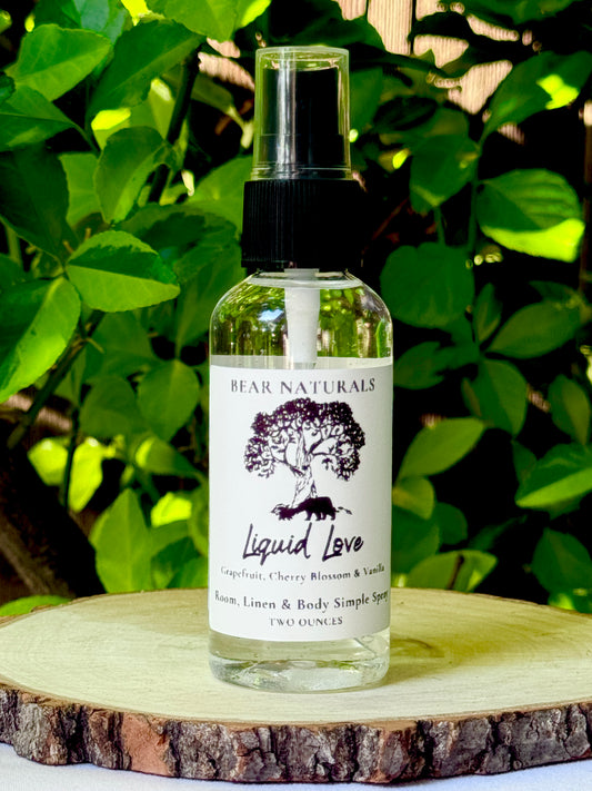 A clear bottle with a black fingertip style spray nozzle featuring a white label with black writing on a wooden surface with green leaves in the background.