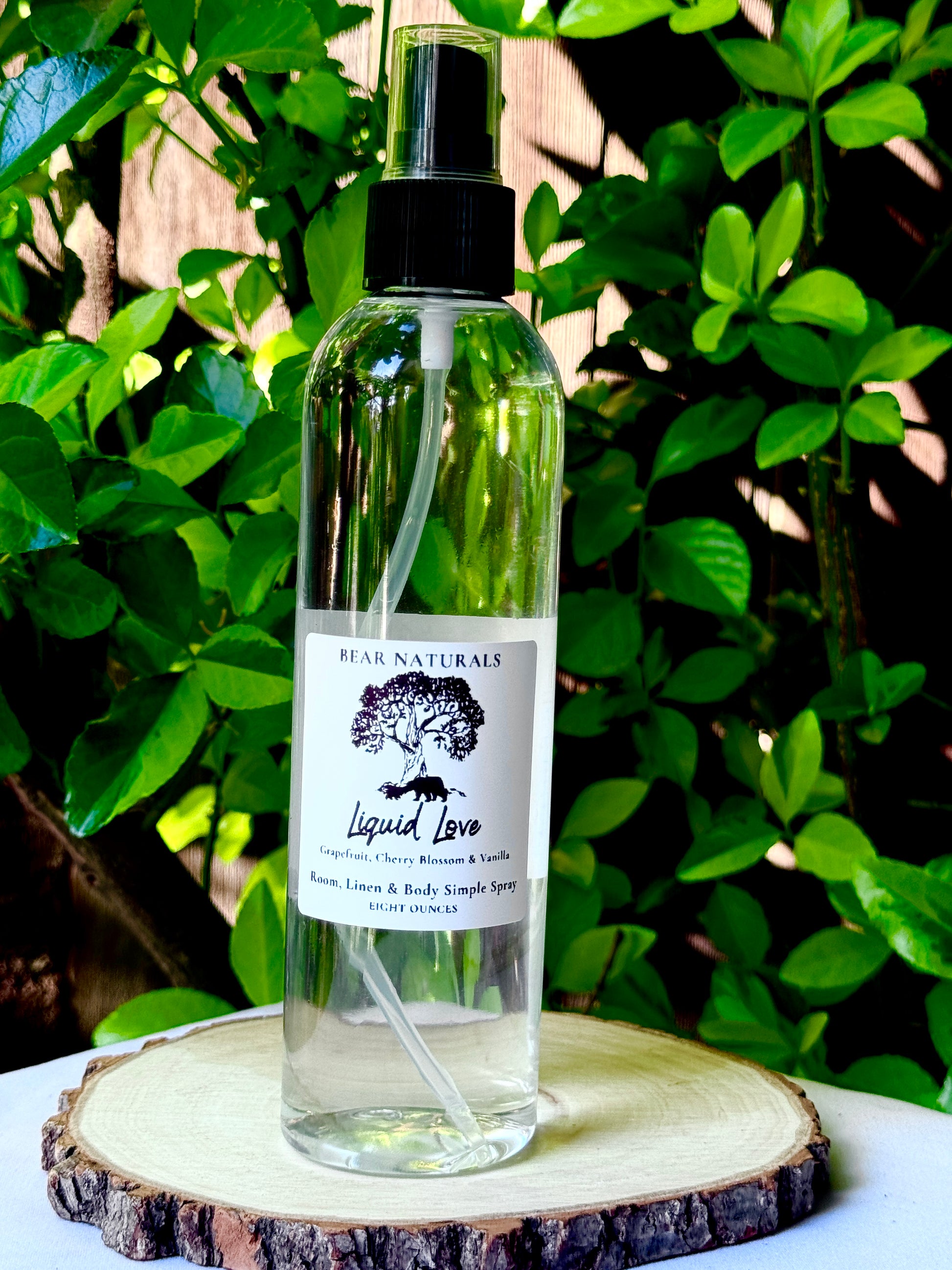 A clear bottle with a black fingertip style spray nozzle featuring a white label with black writing on a wooden surface with green leaves in the background.