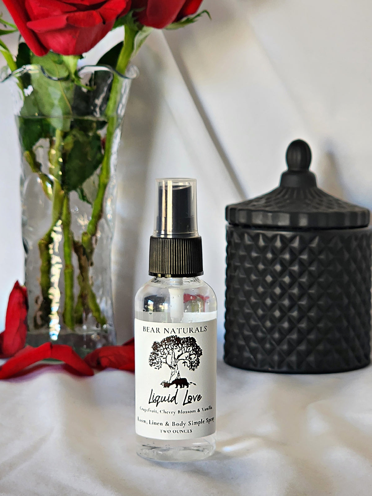 A clear bottle with a black fingertip style spray nozzle featuring a white label with black writing with a black candle jar and red roses in a vase in the background.