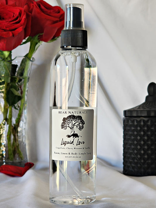 A clear bottle with a black fingertip style spray nozzle featuring a white label with black writing with a black candle jar and red roses in a vase in the background.