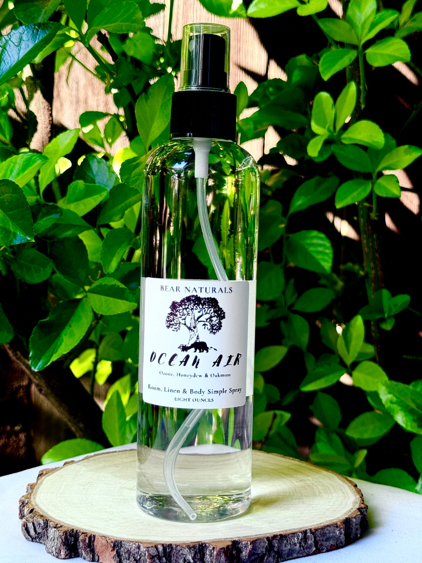 A clear bottle with a black fingertip style spray nozzle featuring a white label with black writing on a wooden surface with green leaves in the background.