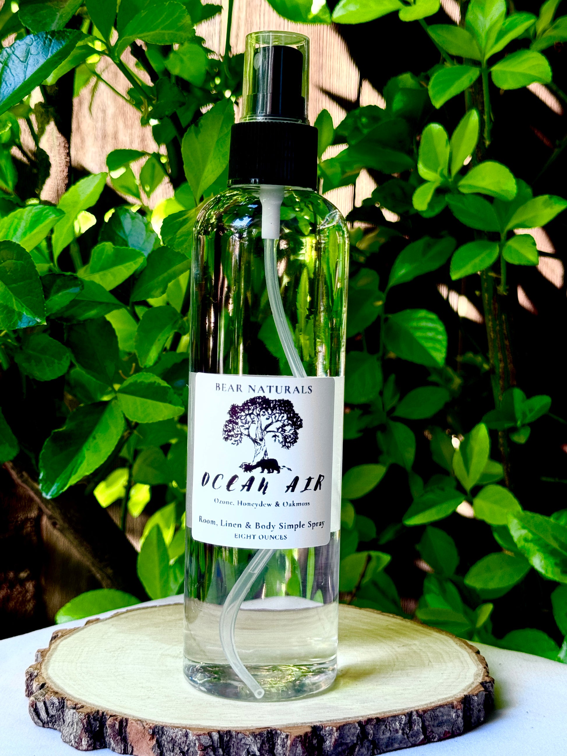 A clear bottle with a black fingertip style spray nozzle featuring a white label with black writing on a wooden surface with green leaves in the background.