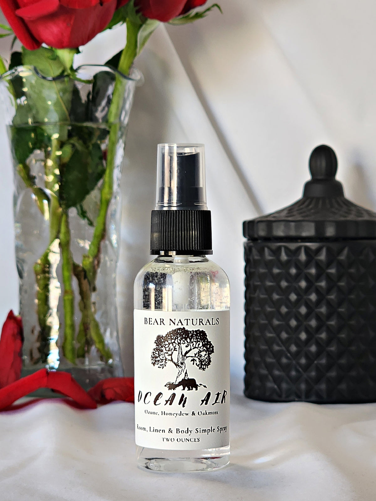 A clear bottle with a black fingertip style spray nozzle featuring a white label with black writing with a black candle jar and red roses in a vase in the background.