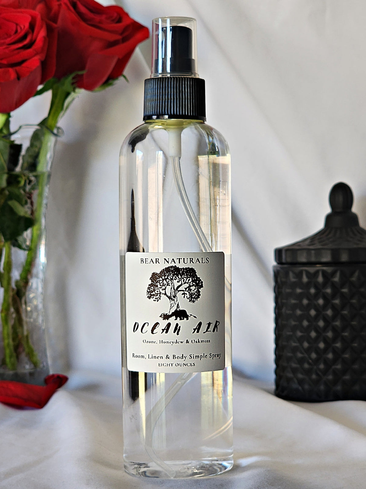 A clear bottle with a black fingertip style spray nozzle featuring a white label with black writing with a black candle jar and red roses in a vase in the background.