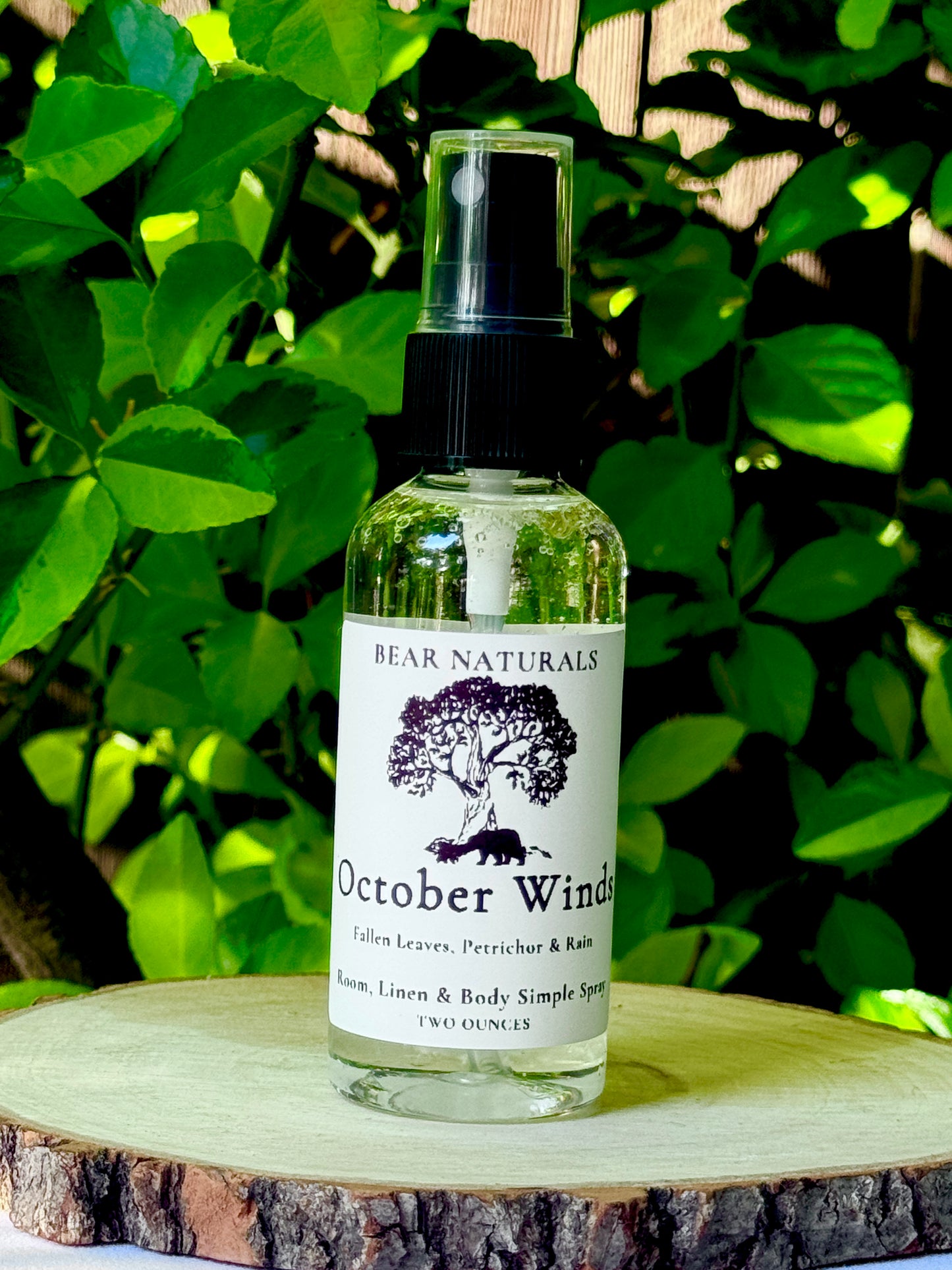 A clear bottle with a black fingertip style spray nozzle featuring a white label with black writing on a wooden surface with green leaves in the background.