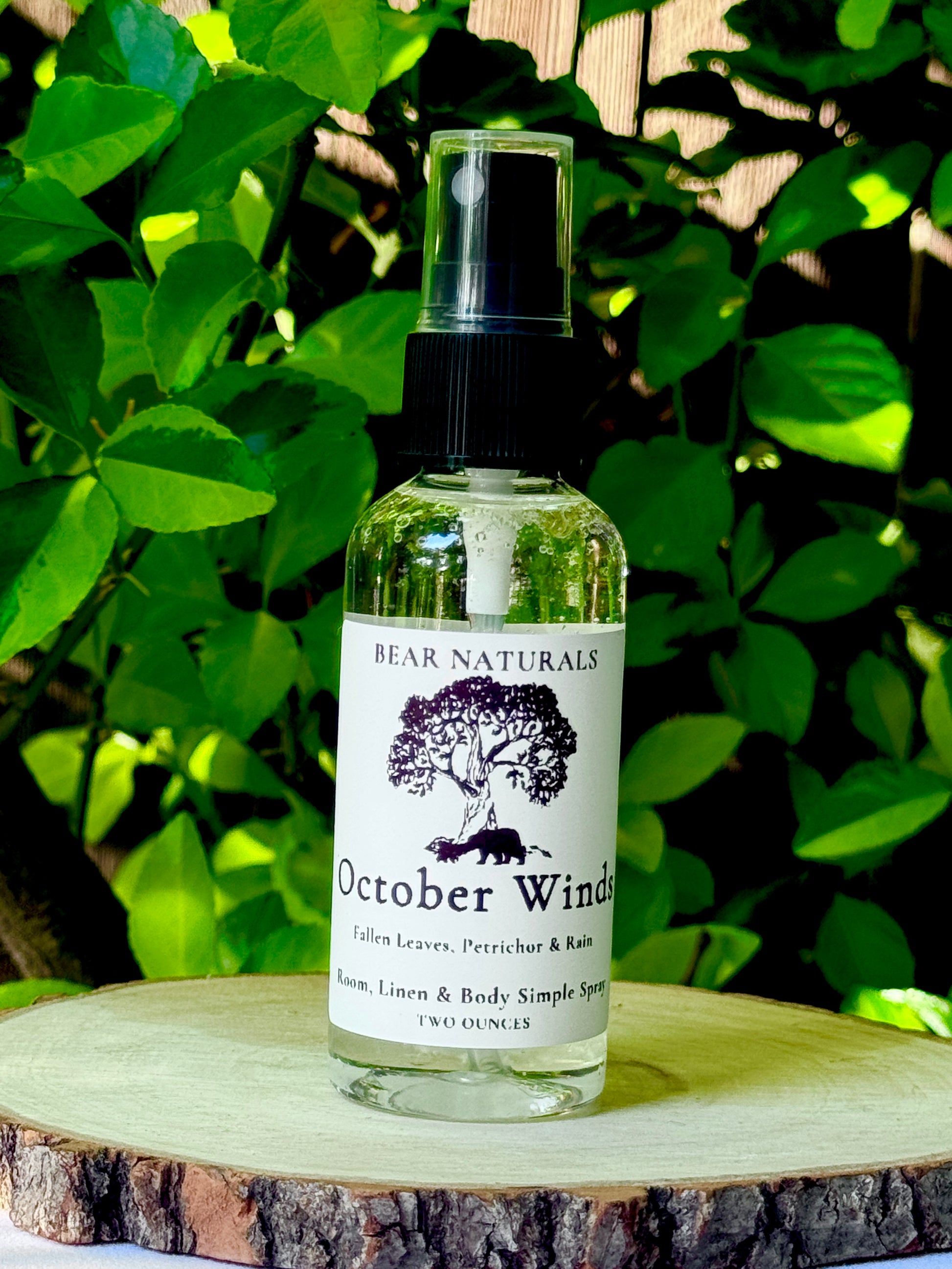 A clear bottle with a black fingertip style spray nozzle featuring a white label with black writing on a wooden surface with green leaves in the background.