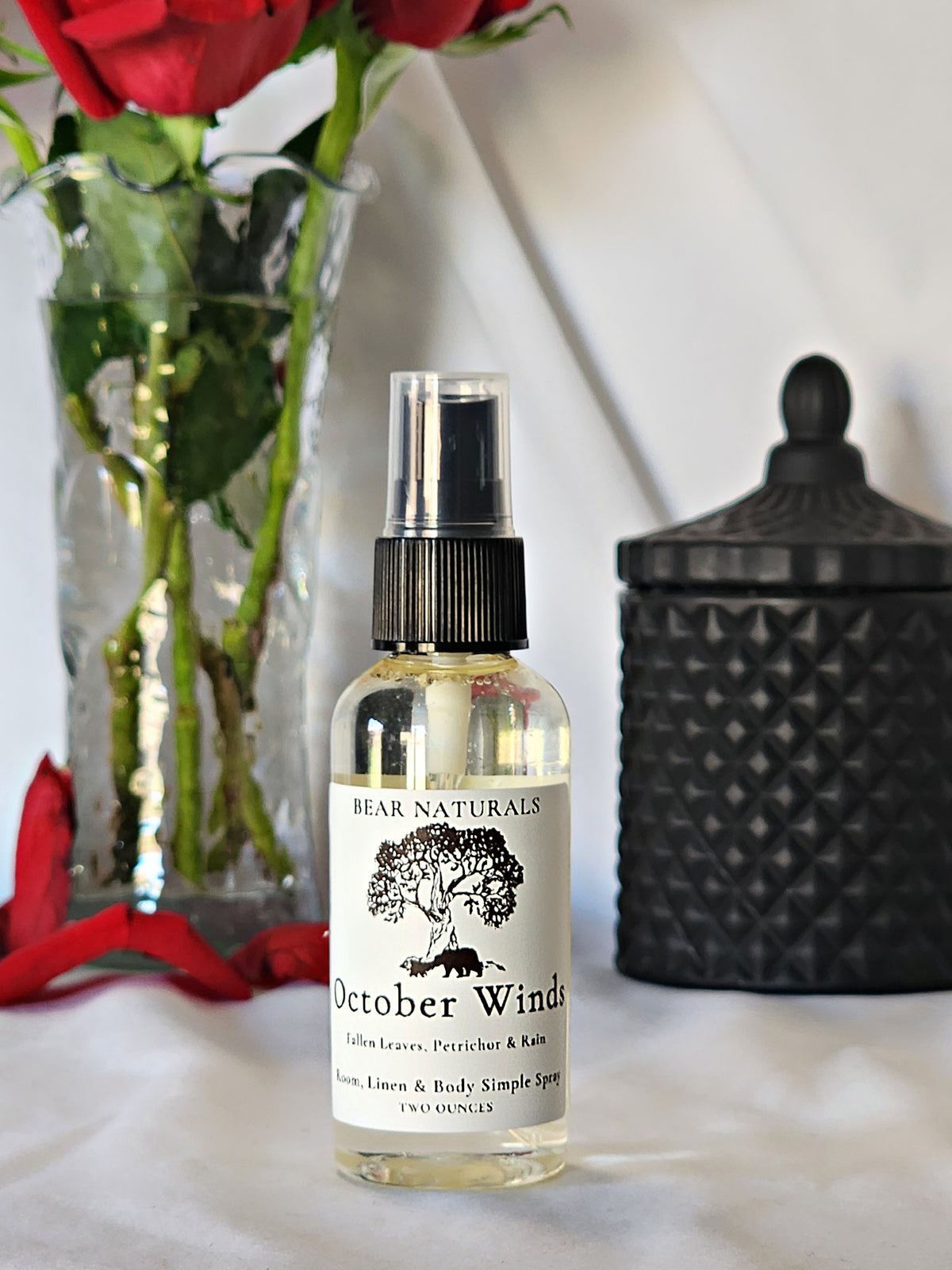 A clear bottle with a black fingertip style spray nozzle featuring a white label with black writing with a black candle jar and red roses in a vase in the background.