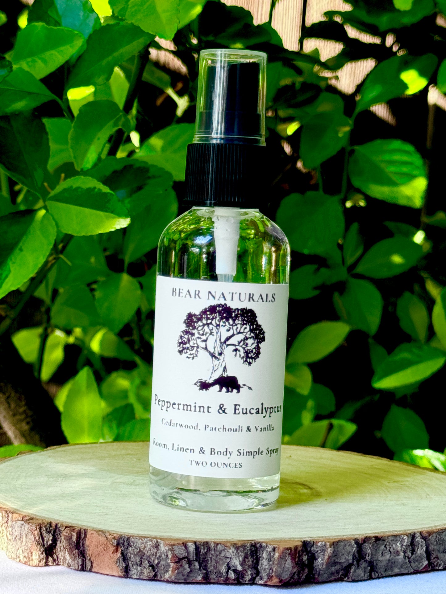 A clear bottle with a black fingertip style spray nozzle featuring a white label with black writing on a wooden surface with green leaves in the background.