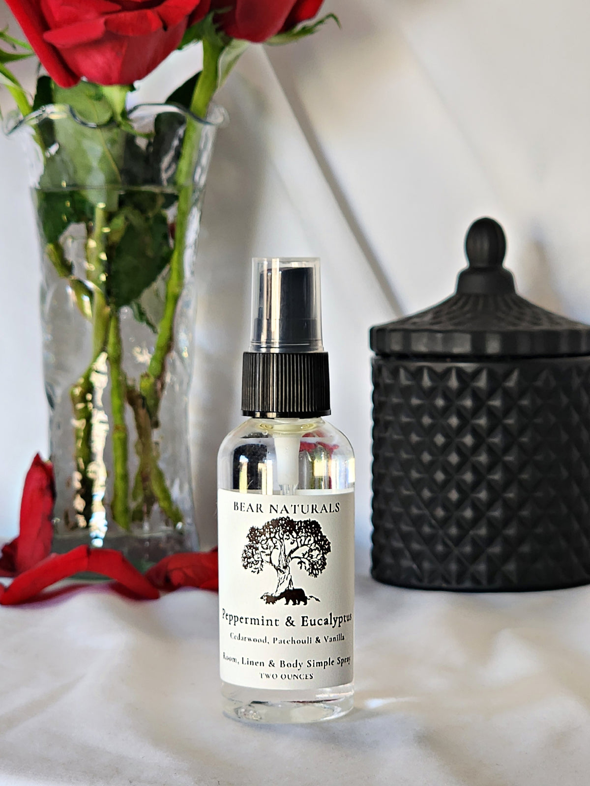 A clear bottle with a black fingertip style spray nozzle featuring a white label with black writing with a black candle jar and red roses in a vase in the background.