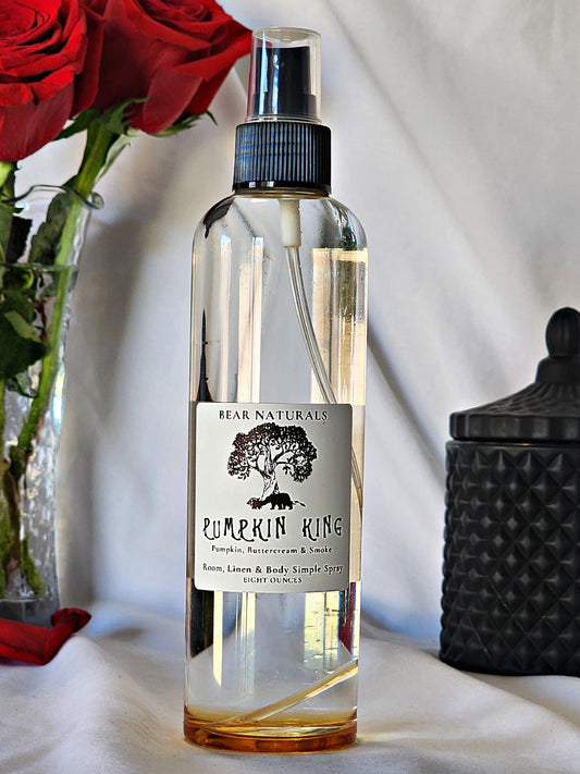 A clear bottle with a black fingertip style spray nozzle featuring a white label with black writing with a black candle jar and red roses in a vase in the background.