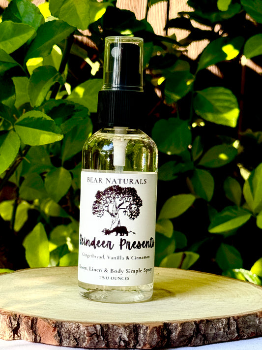 A clear two ounce finger-tip spray bottle with black and white label on a wooden surface with green leaves in the background. The bottle contains the scent "Reindeer Presents".
