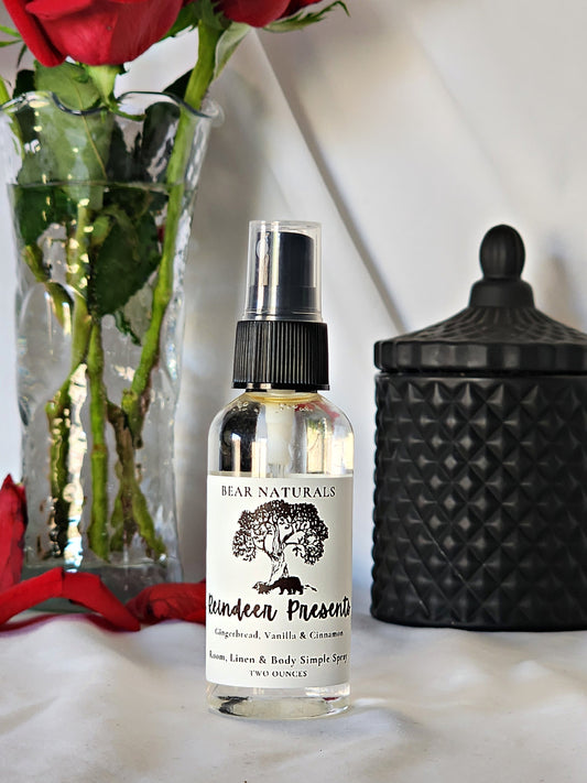 A clear bottle with a black fingertip style spray nozzle featuring a white label with black writing with a black candle jar and red roses in a vase in the background.