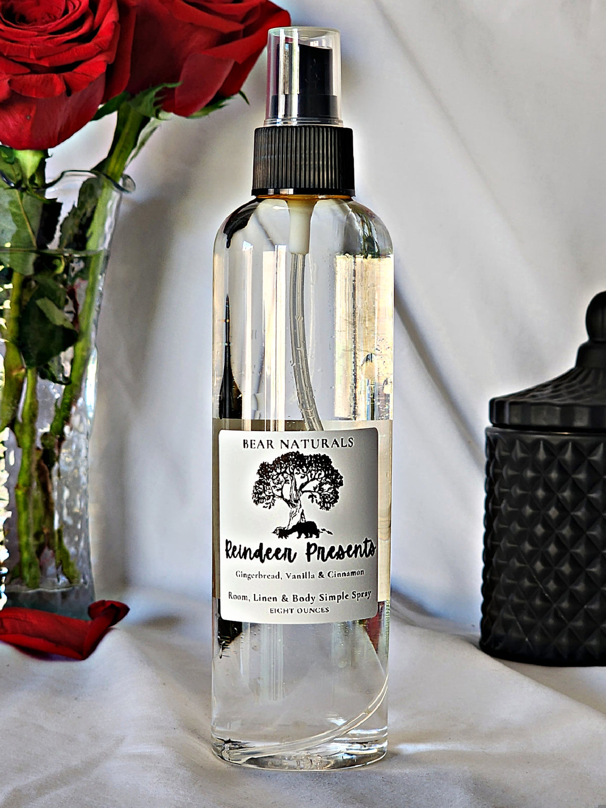 A clear bottle with a black fingertip style spray nozzle featuring a white label with black writing with a black candle jar and red roses in a vase in the background.