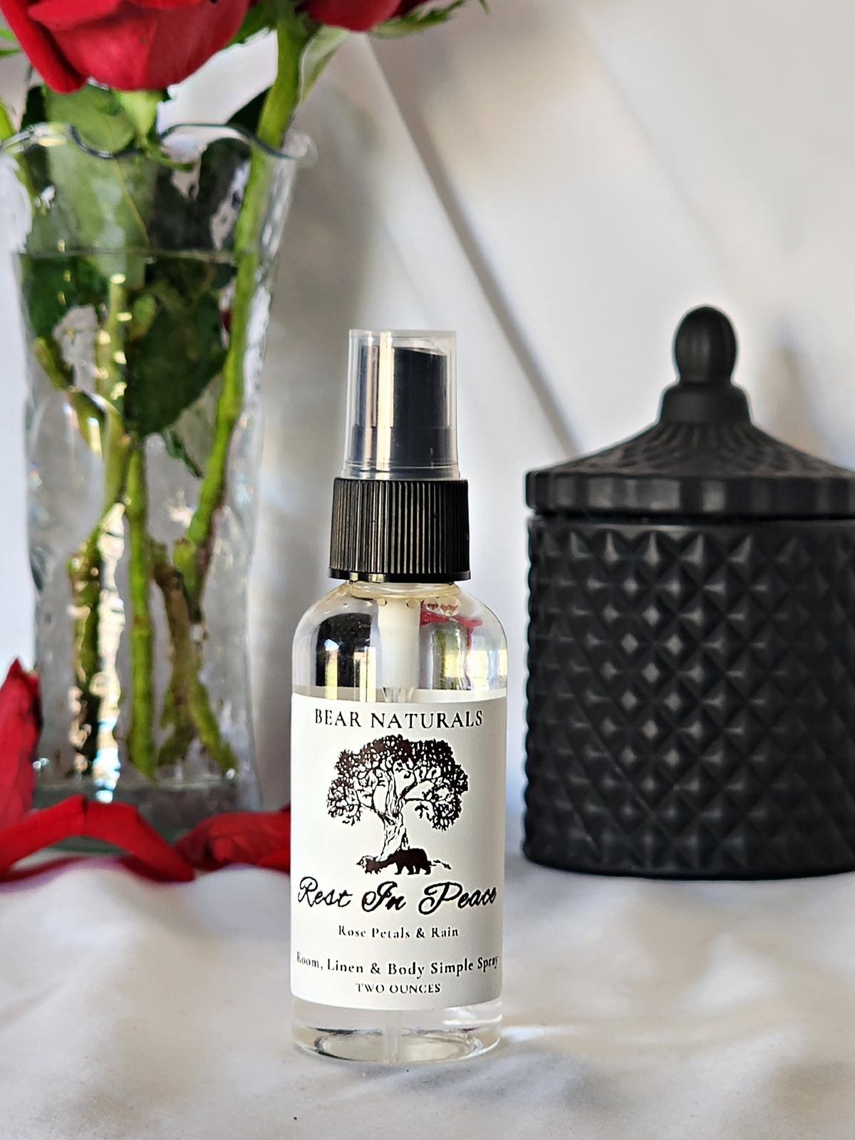 A clear bottle with a black fingertip style spray nozzle featuring a white label with black writing with a black candle jar and red roses in a vase in the background.