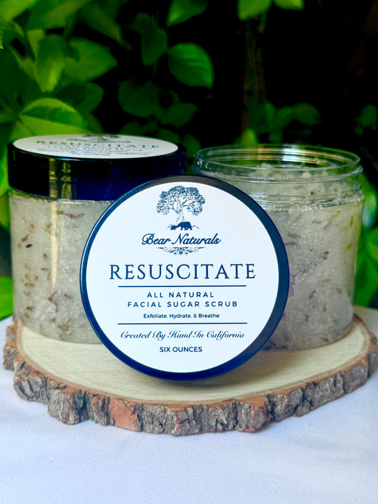 Two clear six ounce plastic jars with black lids featuring a white label with black writing that reads "Resuscitate All Natural Facial Sugar Scrub" filled with product sitting on a wooden base with green leaves in the background..
