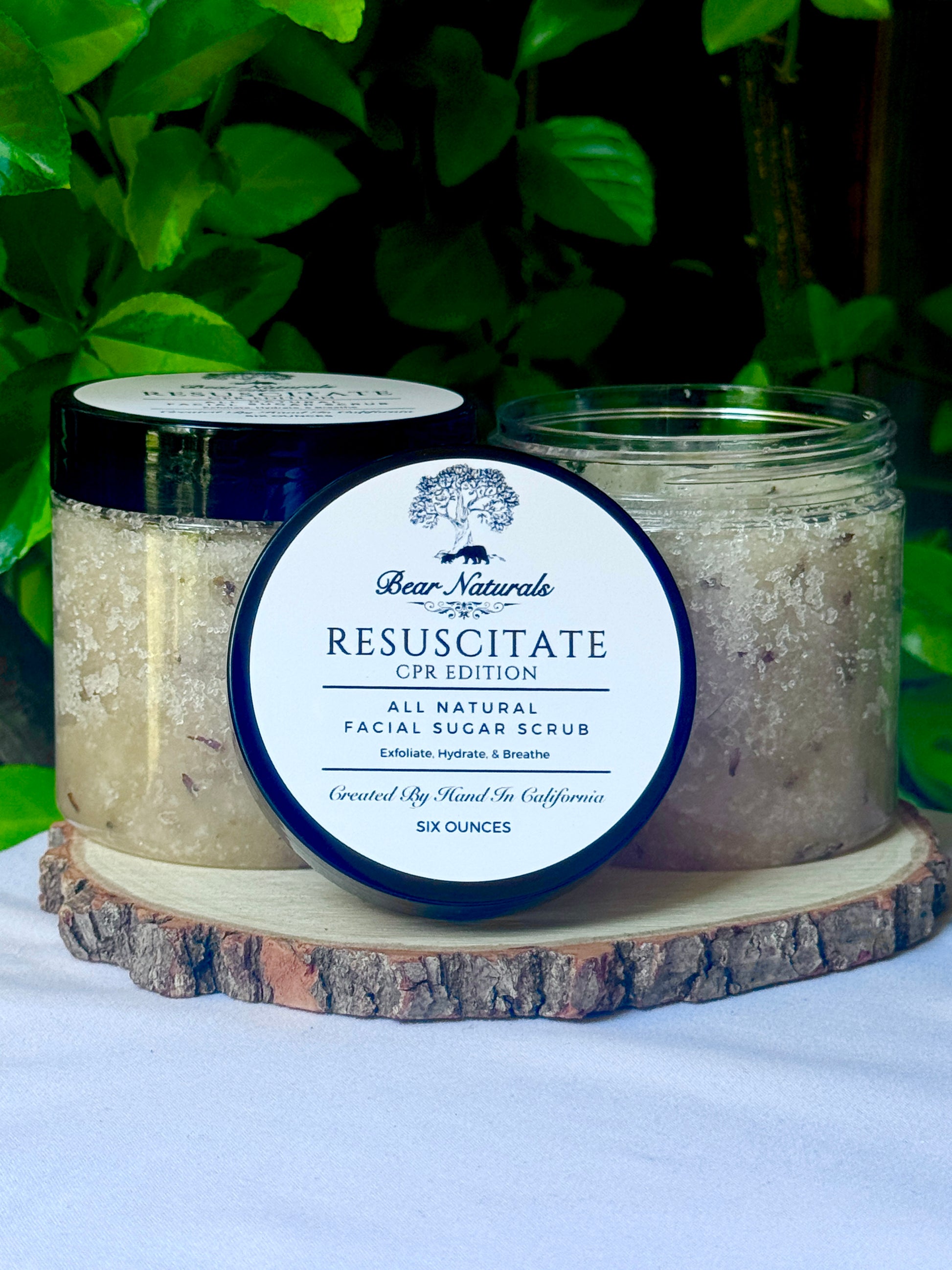 two clear 8 ounce jars of Resuscitate All Natural Exfoliating Sugar Scrub CPR Edition on a wood stand with leaves in the background.