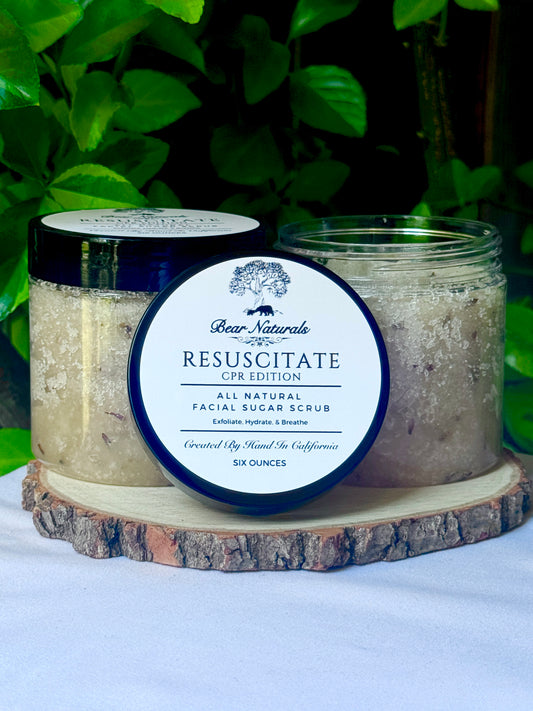 two clear 8 ounce jars of Resuscitate All Natural Exfoliating Sugar Scrub CPR Edition on a wood stand with leaves in the background.